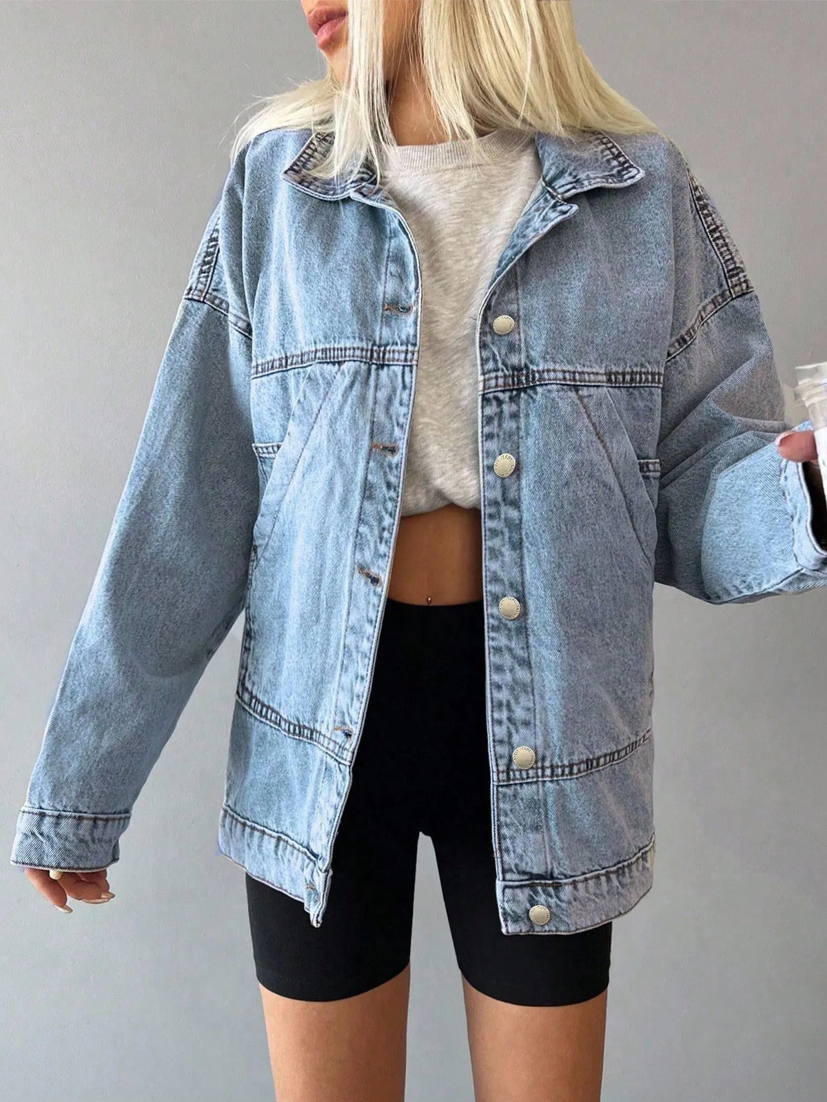 Woman wearing the oversized light-wash denim jacket with a white top and black bike shorts, holding a drink