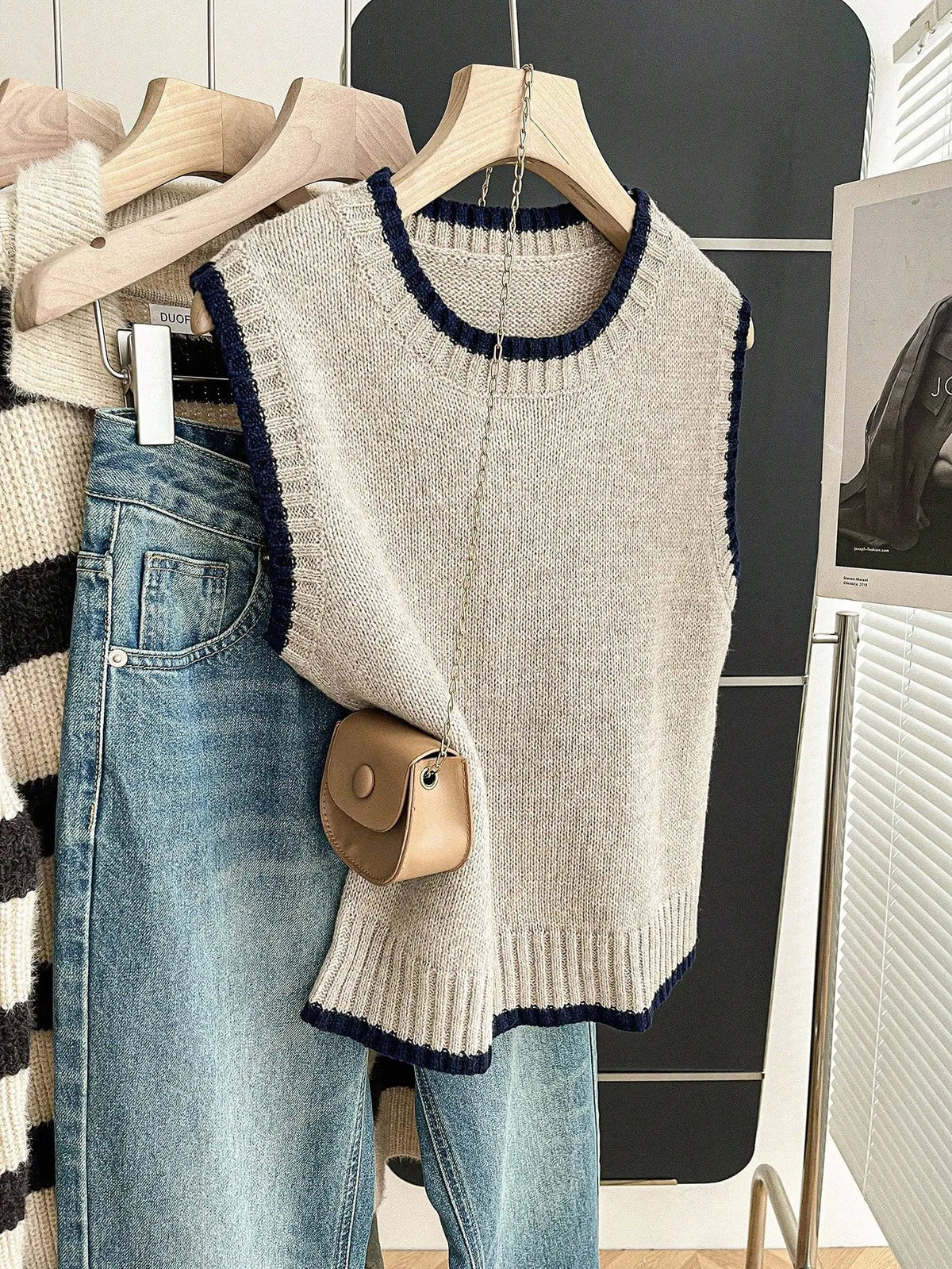 Grey knit sweater vest with navy trim on a hanger beside jeans and a small beige bag