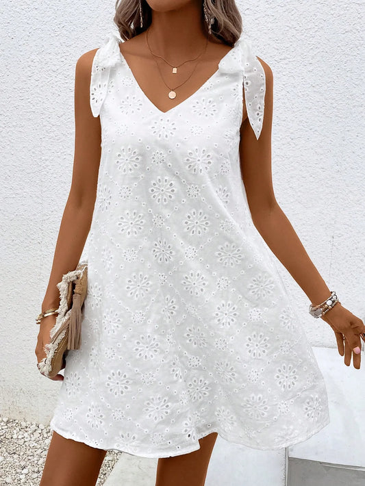 Woman wearing a white eyelet V-neck mini dress with tied shoulder straps, front view with a clutch