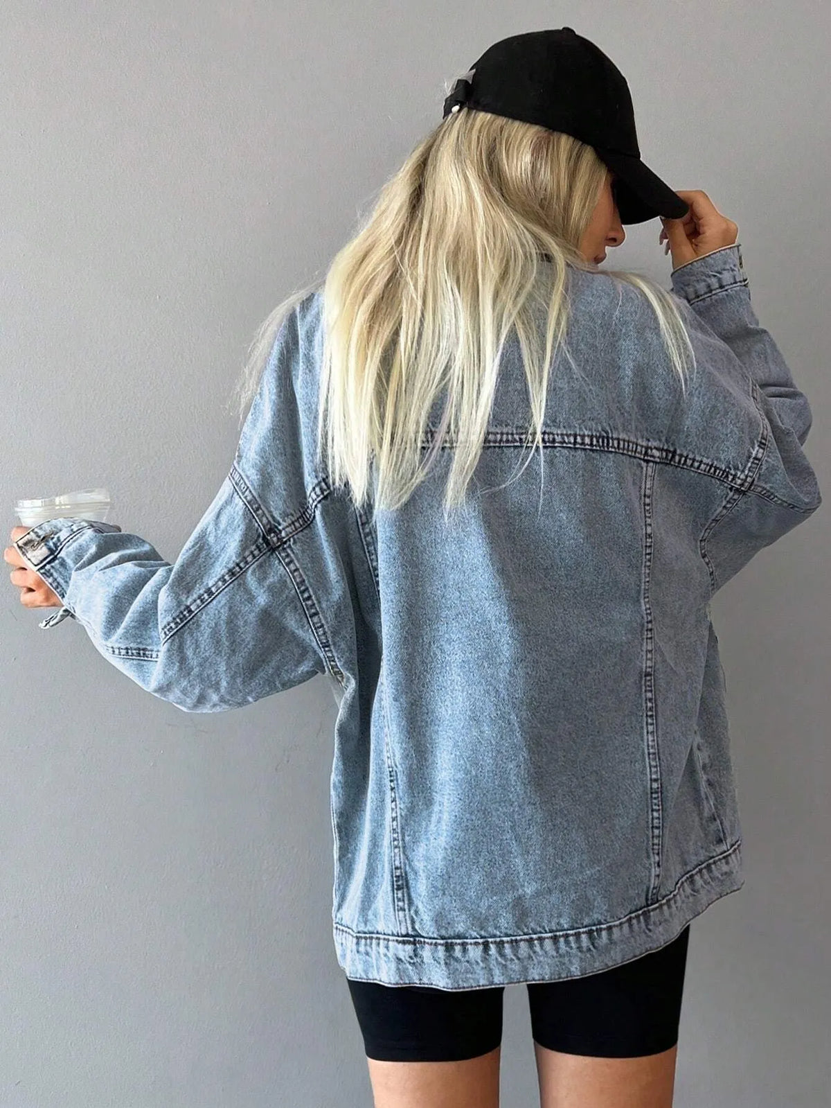 Back view of the oversized light-wash denim jacket worn with a black cap and black bike shorts