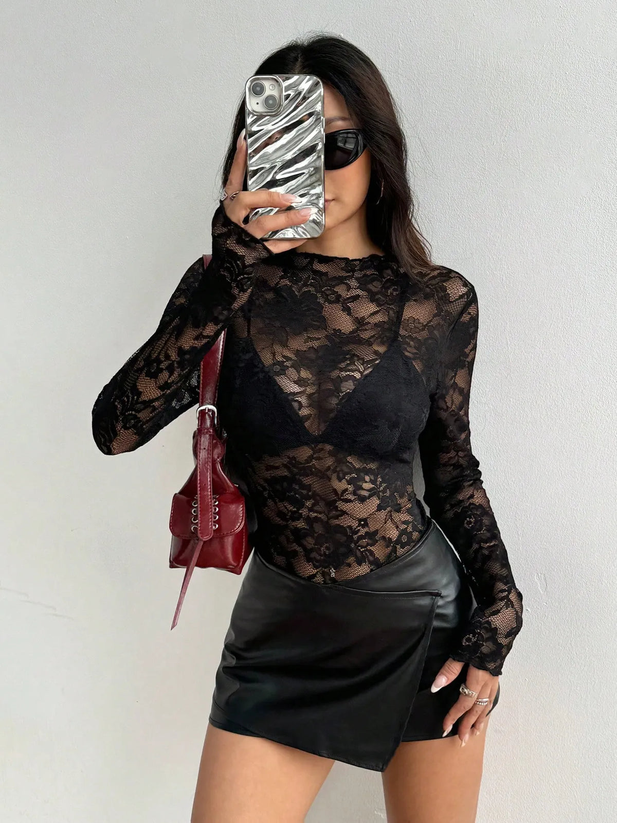 Woman in a black sheer lace long-sleeve top over a black bra with a black mini skirt, mirror selfie holding a red bag