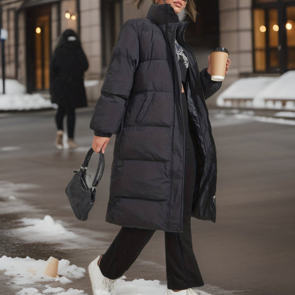 Woman wearing a long black quilted puffer jacket with a high collar, holding a coffee cup and a bag on a snowy street