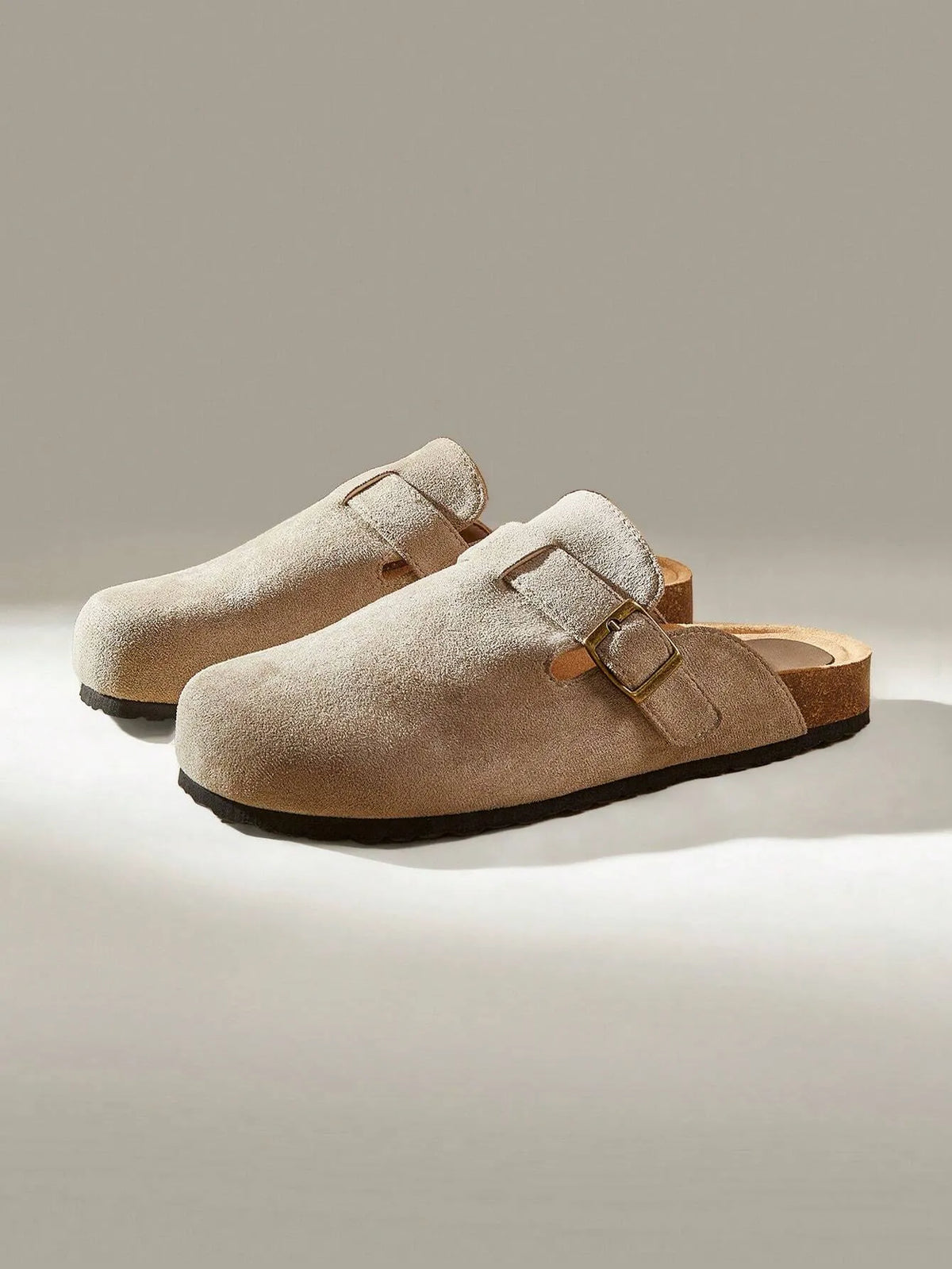 Three-quarter view of a taupe suede clog-style slide with a buckle strap