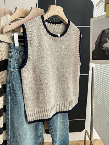Grey knit sweater vest with navy trim at the neck and armholes hanging on a hanger beside jeans