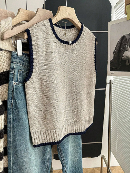 Grey knit sweater vest with navy trim at the neck and armholes hanging on a hanger beside jeans