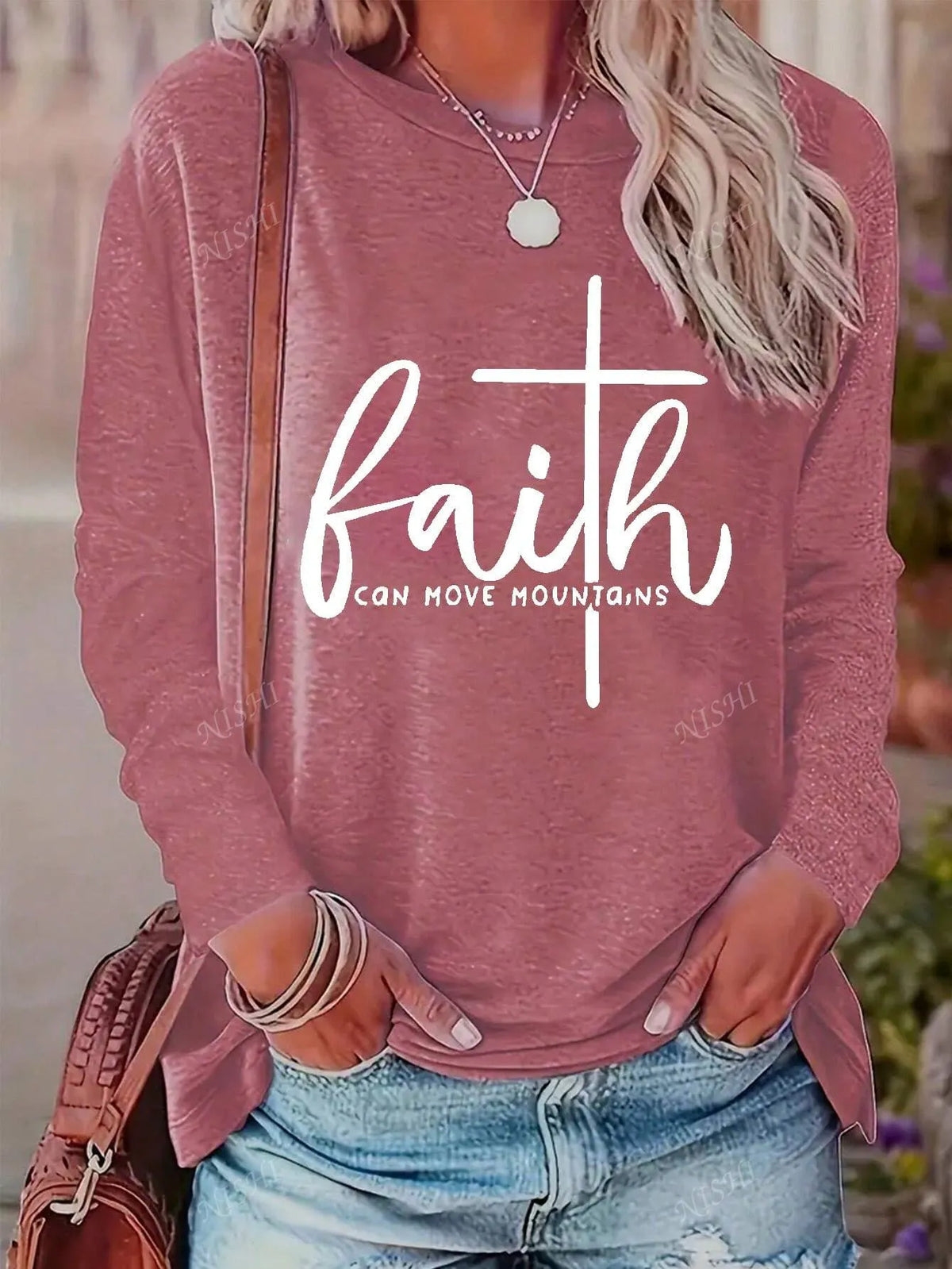 Woman wearing a coral pink long-sleeve tee with a white faith cross graphic and a brown shoulder bag