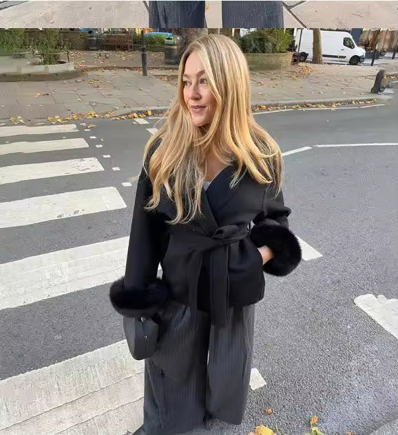 Woman wearing a black wrap coat with a tie belt and fur-trimmed cuffs on a city street