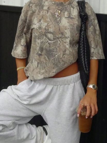 Woman wearing an oversized olive camo print T-shirt with light grey sweatpants and holding a drink, cropped view