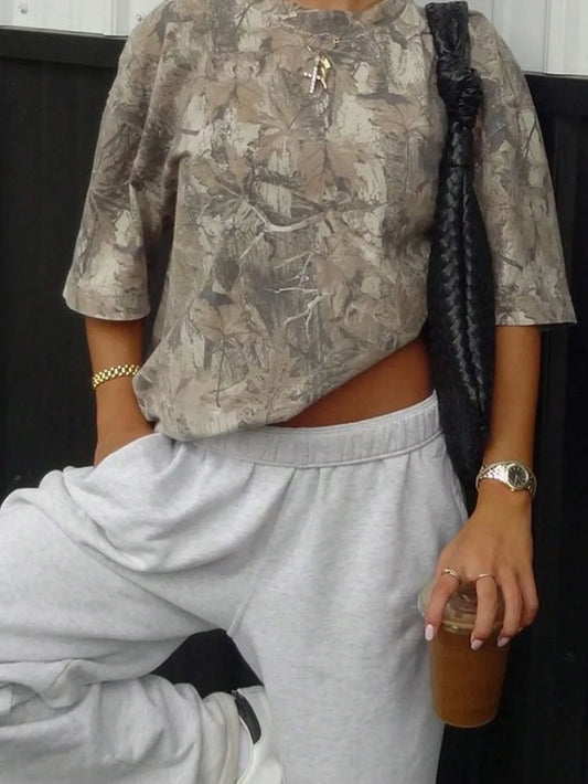 Woman wearing an oversized olive camo print T-shirt with light grey sweatpants and holding a drink, cropped view