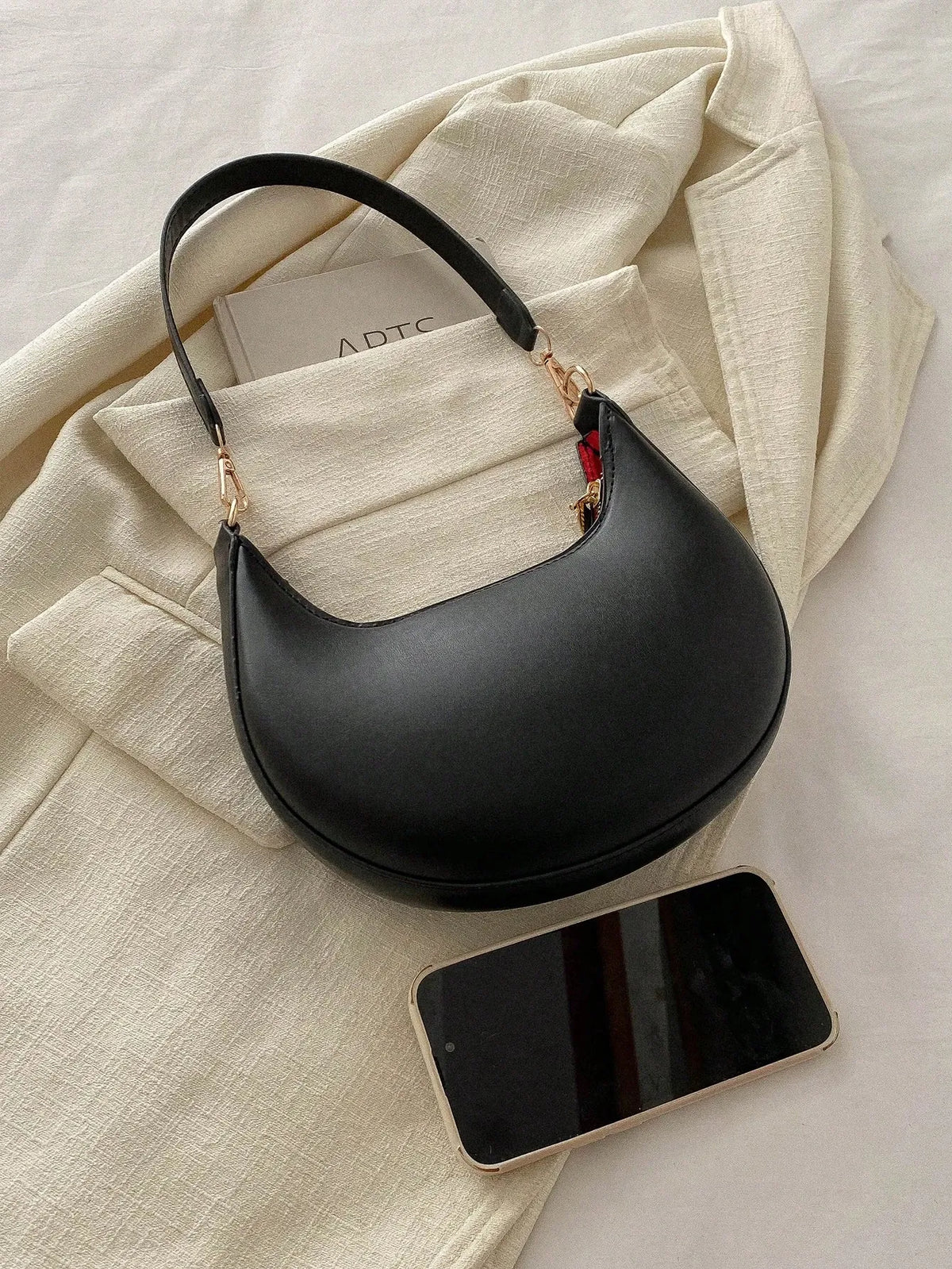 Black half-moon shoulder bag with a looped strap, laid on a cream cloth beside a phone