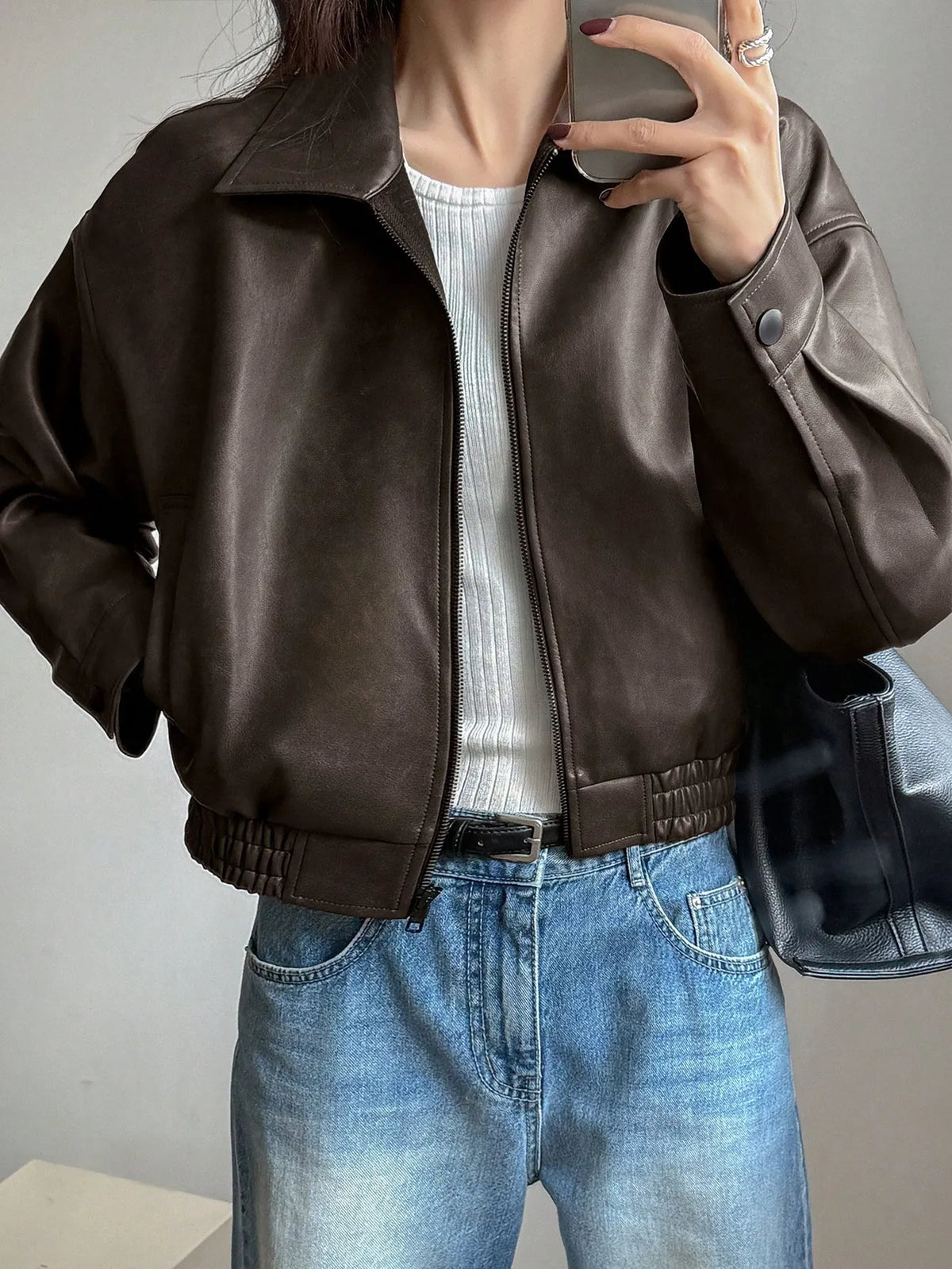 Woman in a cropped dark brown leather-look jacket over a white top and light-wash jeans, taking a mirror selfie
