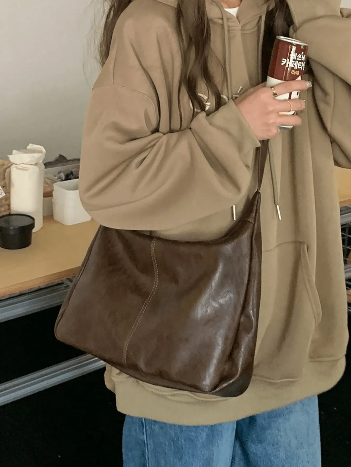 Woman in a tan zip jacket and jeans carrying the brown slouchy bag, holding a cup