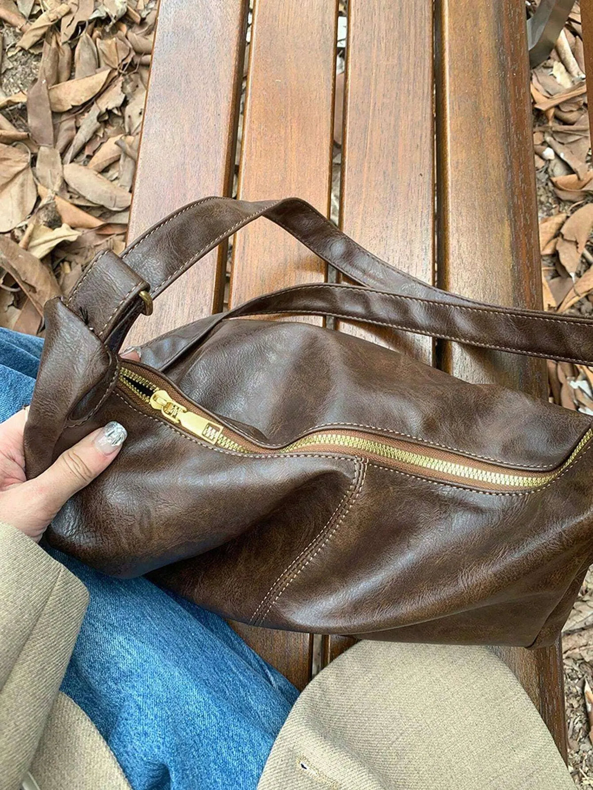 Brown slouchy bag with a gold zipper and two handles, shown on a wooden bench