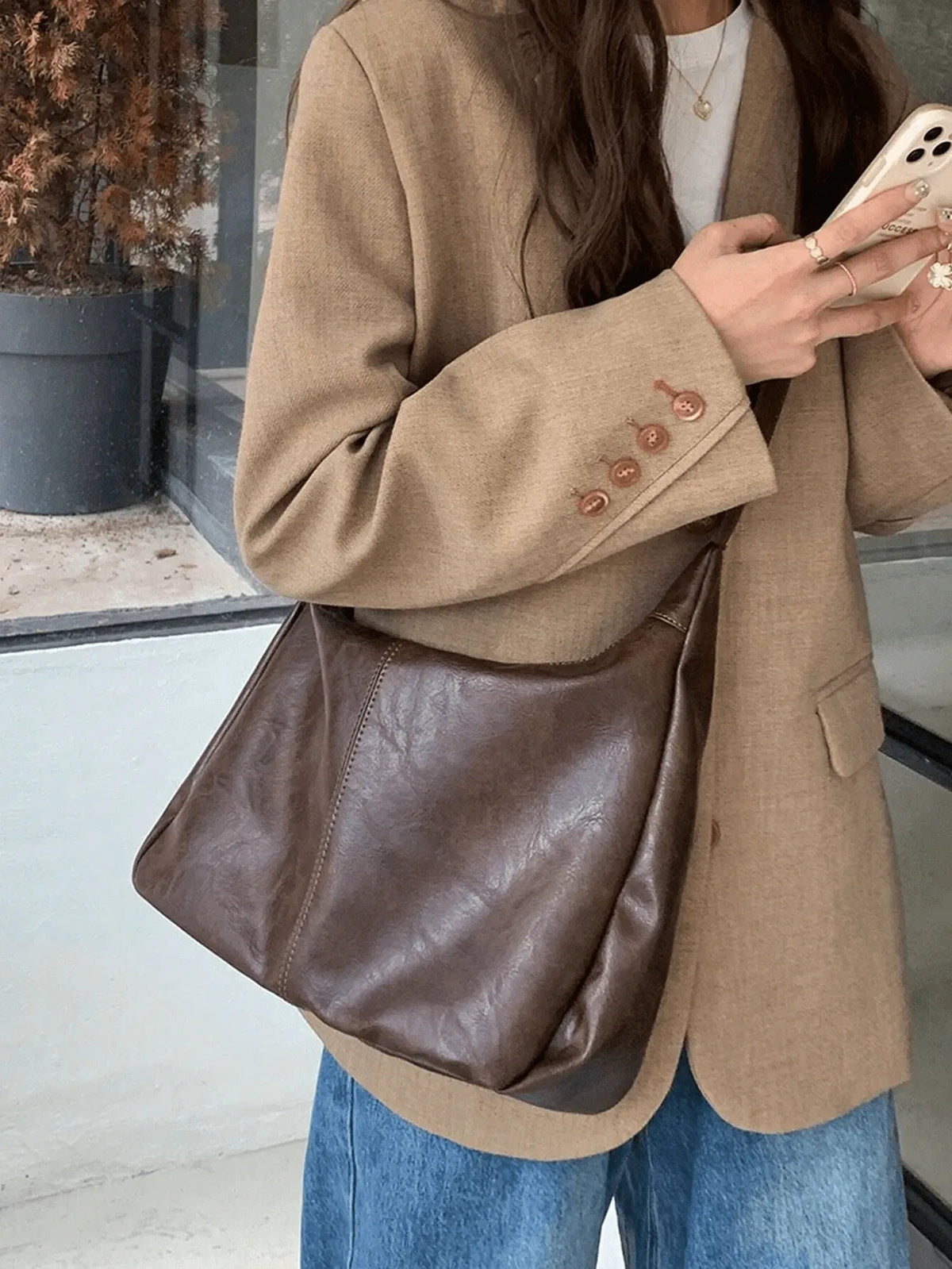 Woman in a tan blazer and jeans carrying a brown slouchy crossbody bag, holding a phone