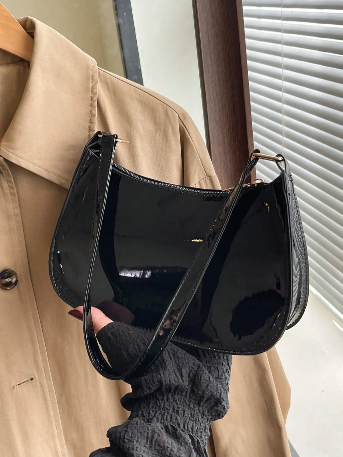 Glossy black half-moon shoulder bag held by its strap against a beige jacket