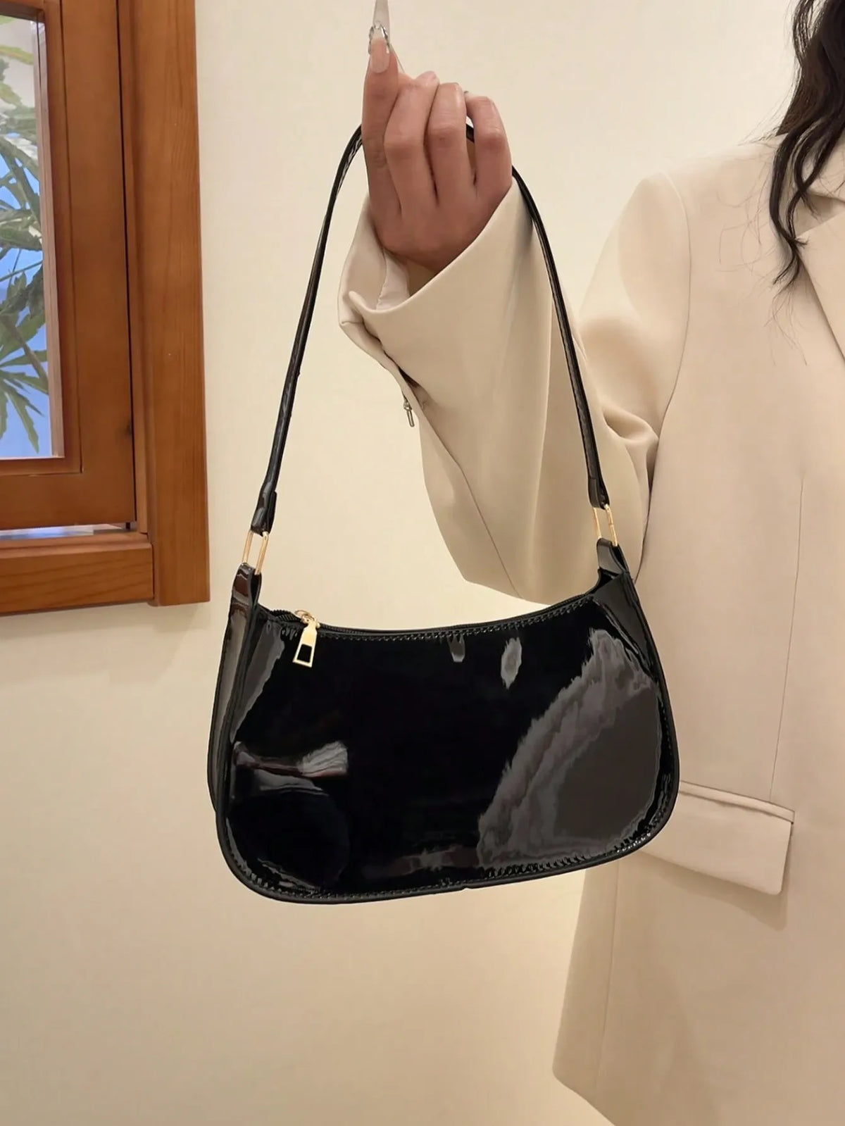 Glossy black half-moon shoulder bag held up by the strap, worn with a white blazer