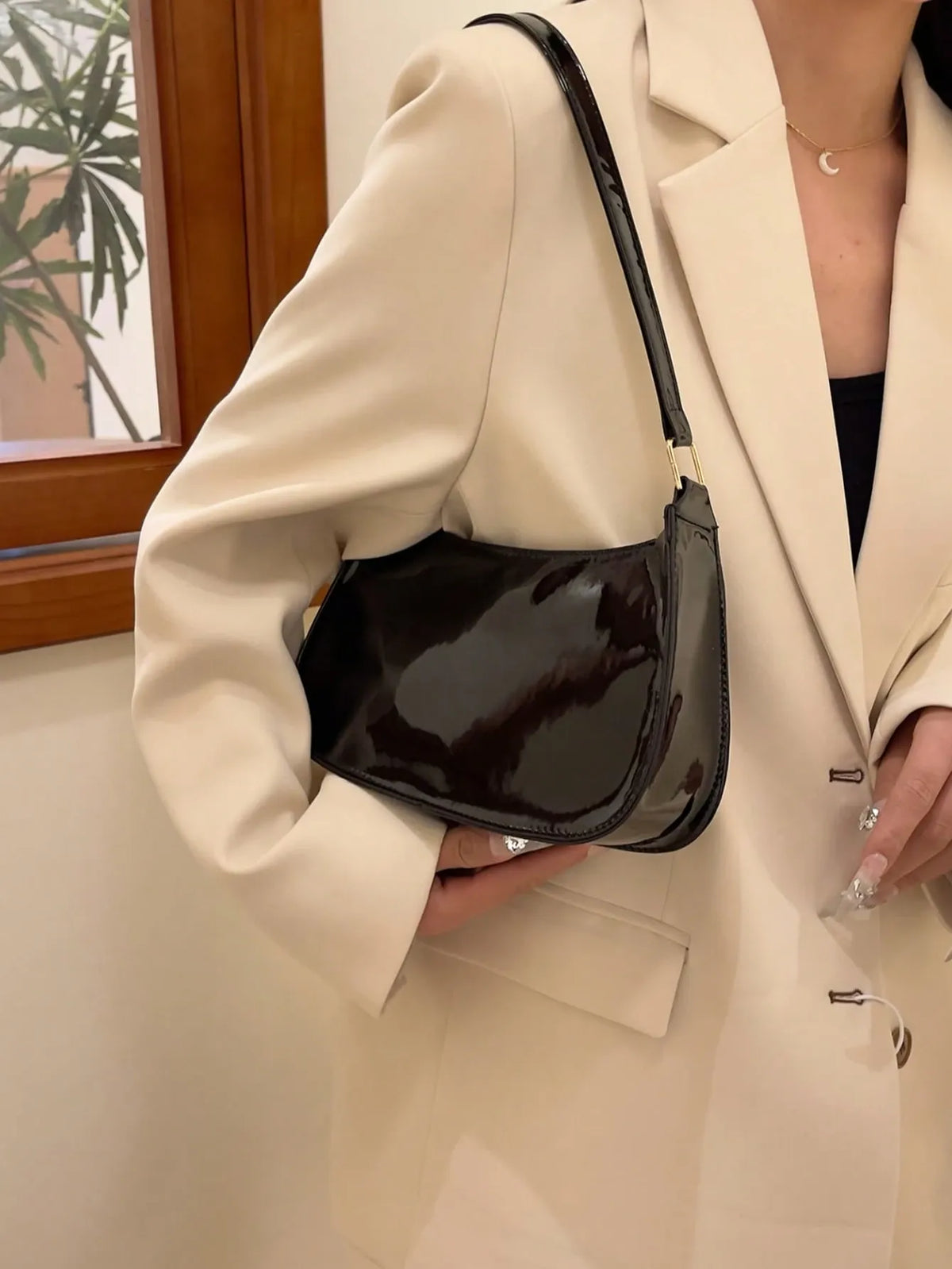 Glossy black half-moon bag carried at her side with a beige blazer, close view