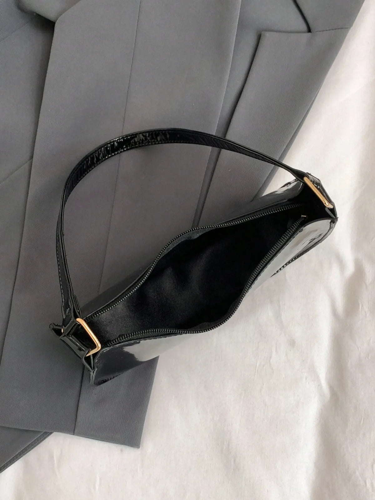 Glossy black half-moon bag with a gold buckle detail, laid flat on grey fabric