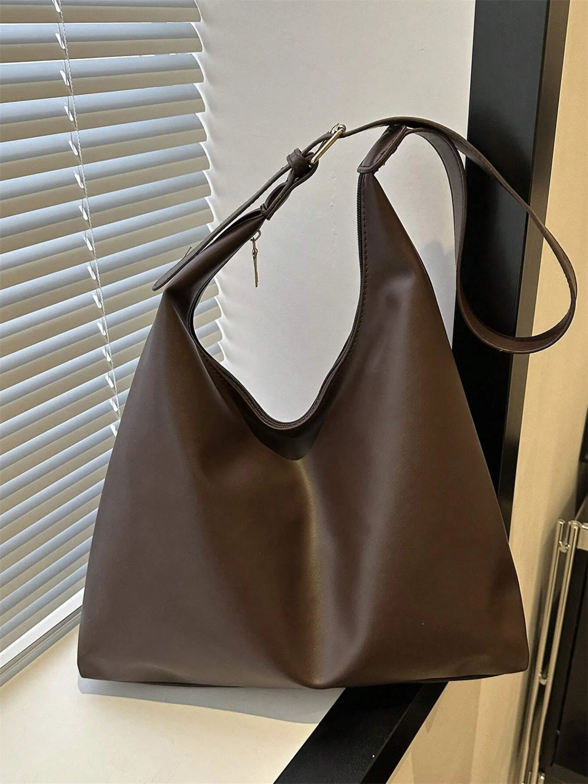 Dark brown shoulder bag with the strap raised, standing by a window